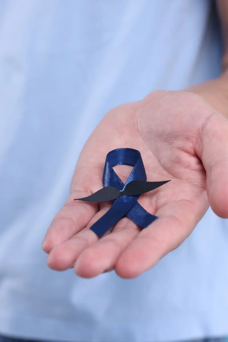 Prostate cancer awareness. Man holding blue ribbon with fake mustache on color background, closeup